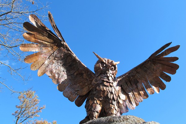 copper sculpture of owl with wings spread outside in front of trees