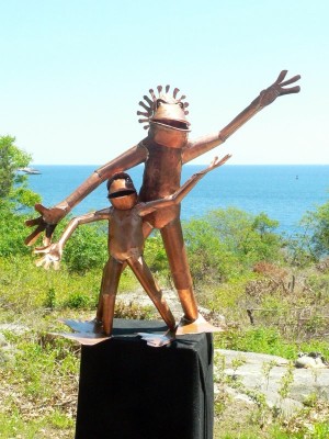 Copper Sculpture of large frog with crown standing next to small frog with arms extended on wooden stump in green field in front of ocean