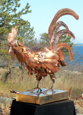 copper rooster sculpture with large tail feathers on pedestal outside in green field in front of water